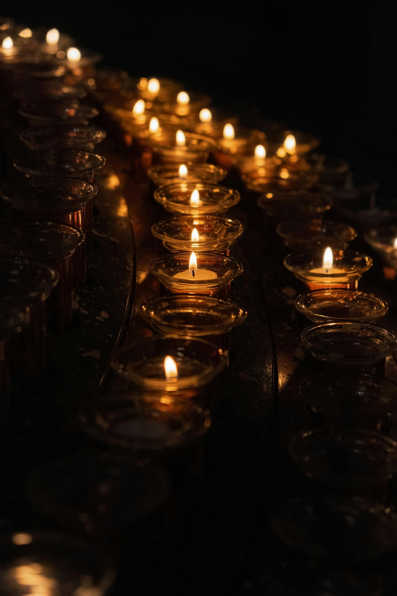 Three rows of votive candles in the dark with some of them still burning
